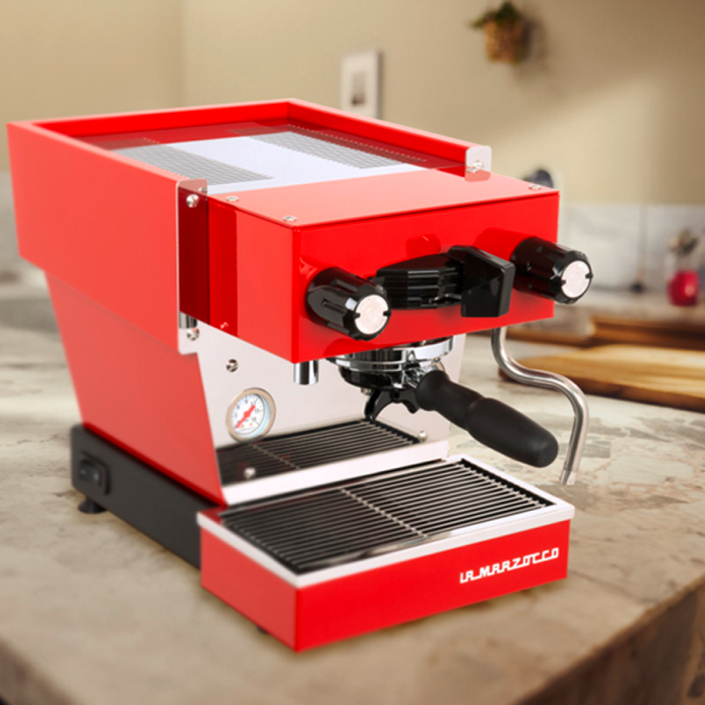 Eine rote Espressomaschine von La Marzocco steht auf einer Küchenarbeitsplatte und ist mit einem Siebträger, einer Dampfdüse, einem Messgerät und einer Metalltropfschale ausgestattet.