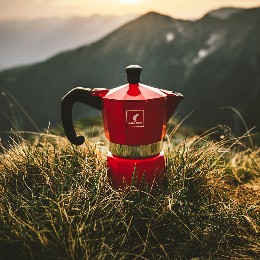 Eine rote Kaffeemaschine steht auf einer Wiese mit Bergen im Hintergrund, fotografiert bei Sonnenaufgang oder Sonnenuntergang.