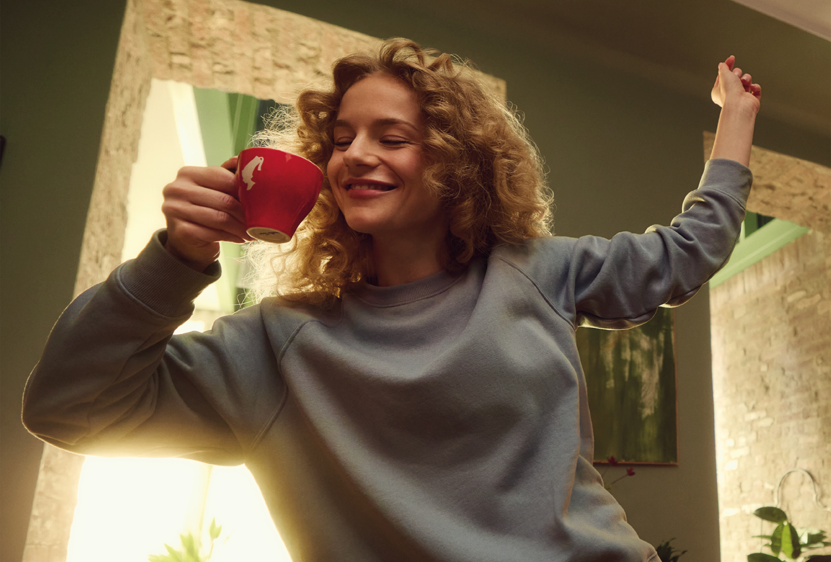 Eine lächelnde Frau mit lockigem Haar genießt eine rote Tasse Kaffee oder Tee und hebt fröhlich einen Arm in einem gemütlichen, sonnenbeschienenen Raum mit Backsteinwänden und grünen Akzenten.